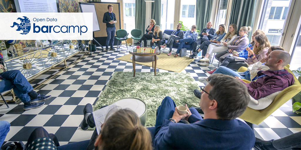 Ein Foto vom Open Data Barcamp 2024. Viele Personen sitzen in einem bunten Raum. Vorne steht eine Person und moderiert eine Session. Links oben auf dem Foto ist das Logo des Open Data Barcamps.