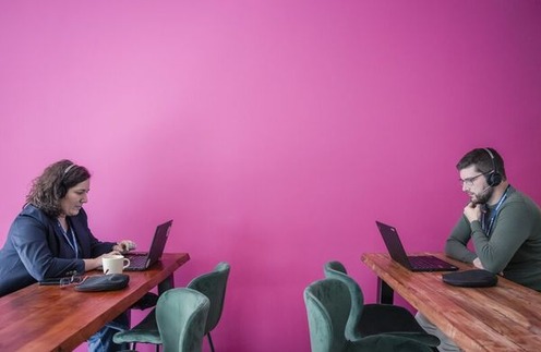Vor einer pinken Wand sitzt links eine Frau an einem Laptop und Tisch. Mit etwas Abstand rechts daneben sitzt ein Mann an Tisch und Laptop und hat Kopfhörer auf.