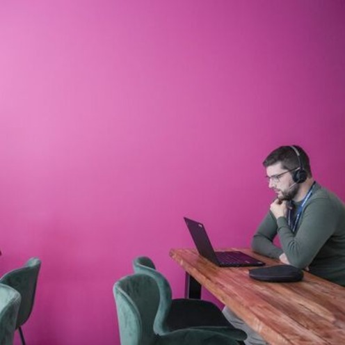 Vor einer pinken Wand sitzt links eine Frau an einem Laptop und Tisch. Mit etwas Abstand rechts daneben sitzt ein Mann an Tisch und Laptop und hat Kopfhörer auf. 