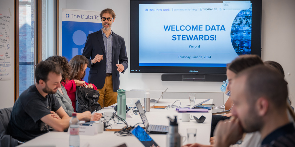 2024_06_13_Data_Stewards_sas_0013 Vorne in einem hell beleuchteten Raum steht Stefaan Verhulst. Neben ihm ein Monitor auf dem steht: Welcome Data Stewards. Im Raum sind einige Teilnehmerinnen und Teilnehmer des Kurses.
